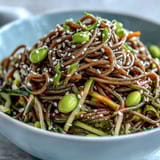 A vibrant Soba Noodle Bowl topped with crisp cucumber ribbons, carrots, and green edamame, drizzled with glossy sesame dressing.